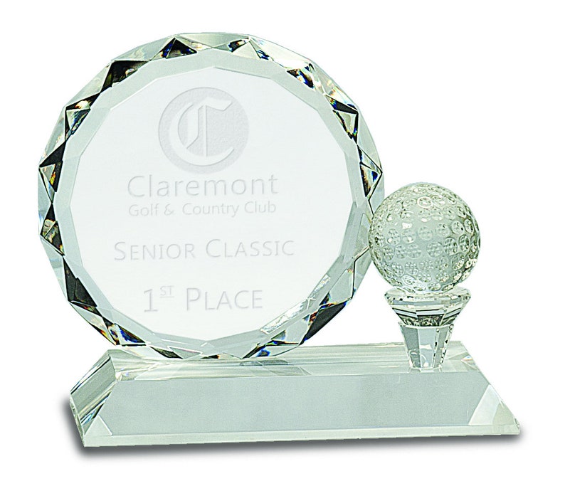 Puede incluir: Un trofeo de golf de cristal transparente con una placa redonda que dice "Claremont Golf & Country Club Senior Classic 1st Place". Una bola de golf se sienta en la parte superior de un tee.