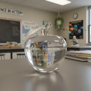 Puede incluir: Un premio transparente en forma de manzana de cristal con un detalle de tallo y hoja. La manzana est&aacute; grabada con las palabras "Mrs. Robert's Earth Science" y "My Pleasant Middle School". El premio est&aacute; sobre un escritorio gris.