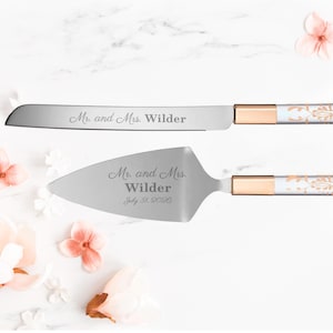 May include: A silver cake knife and server set with gold handles and white floral designs. The knife is engraved with "Mr. and Mrs. Wilder" and the server is engraved with "Mr. and Mrs. Wilder July 31, 2026".