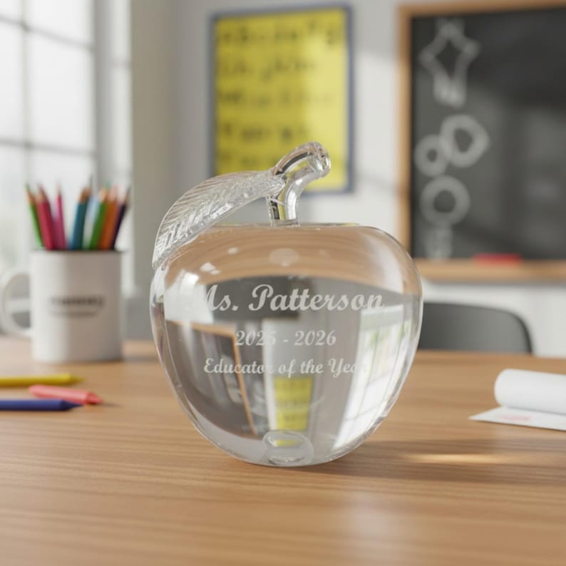 May include: A clear glass apple-shaped award with a leaf-shaped stem. The award is engraved with the name "Ms. Patterson", the years "2025-2026", and the text "Educator of the Year". The award sits on a wooden desk.