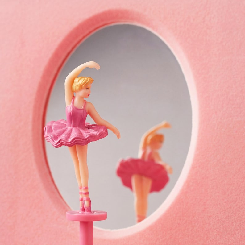 May include: A pink ballerina figurine with a matching tutu and ballet shoes, posed in front of a mirror. The figurine has blonde hair and is set against a pink background. The mirror reflects the ballerina's pose.