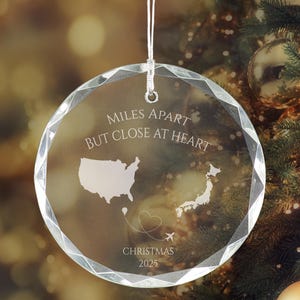 May include: A clear, circular, faceted glass ornament hanging by a white string. It is engraved with the text MILES APART BUT CLOSE AT HEART, maps of the USA and Japan, a dashed flight path forming a heart shape with an airplane, and CHRISTMAS 2025.