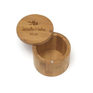 May include: A round, wooden box with a lid. The lid is engraved with the names "Samantha & Andrew" and the year "EST. 2019" along with a decorative leaf design. The box is open, revealing the interior.