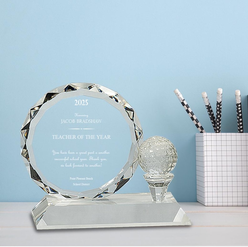 Puede incluir: Un premio de cristal transparente con una bola de golf en la parte superior. El premio dice "2025 Honoring Jacob Bradshaw Teacher of the Year".