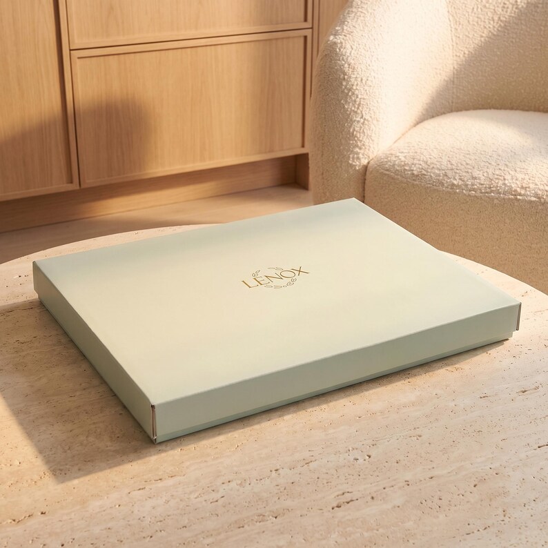 May include: A rectangular, light sage green cardboard box with a flat lid. The center of the lid features the word LENOX printed in gold capital letters, accompanied by a small, delicate gold leaf graphic design positioned above the text.