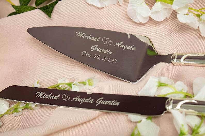 May include: A silver cake cutting set with two knives. The larger knife has the inscription "Michael Angela Guertin Dec. 26, 2020" with two hearts. The smaller knife has the inscription "Michael Angela Guertin" with two hearts.