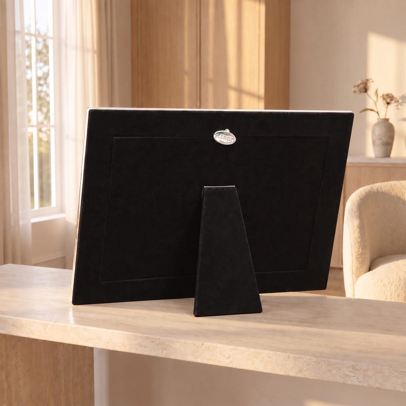 May include: A rectangular picture frame shown from the back, featuring a black velvet-covered backing and a matching easel stand. A small, oval-shaped silver-toned metal plaque with the text "LENOX" is centered near the top edge of the frame.