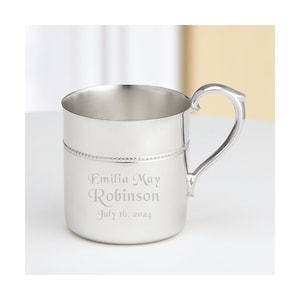 May include: A silver-colored metal mug with a curved handle. The mug has a beaded design around the middle and is engraved with the name "Emilia May Robinson" and the date "July 16, 2024".