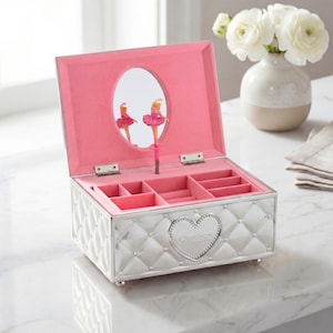 May include: Silver jewellery box with a quilted pattern and a heart-shaped emblem that reads "Charlotte." The interior is lined with pink fabric and features compartments for storage. A mirror reflects two ballerina figurines.