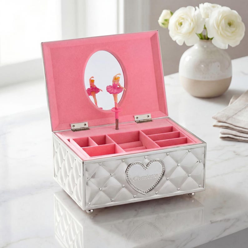 May include: Silver jewellery box with a quilted pattern and a heart-shaped emblem that reads "Charlotte." The interior is lined with pink fabric and features compartments for storage. A mirror reflects two ballerina figurines.