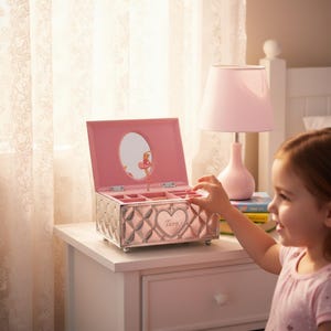 May include: A pink jewelry box with a quilted design and a heart-shaped emblem that reads "Tara." The box is open, revealing compartments and a mirror. A pink lamp sits nearby on a white bedside table.