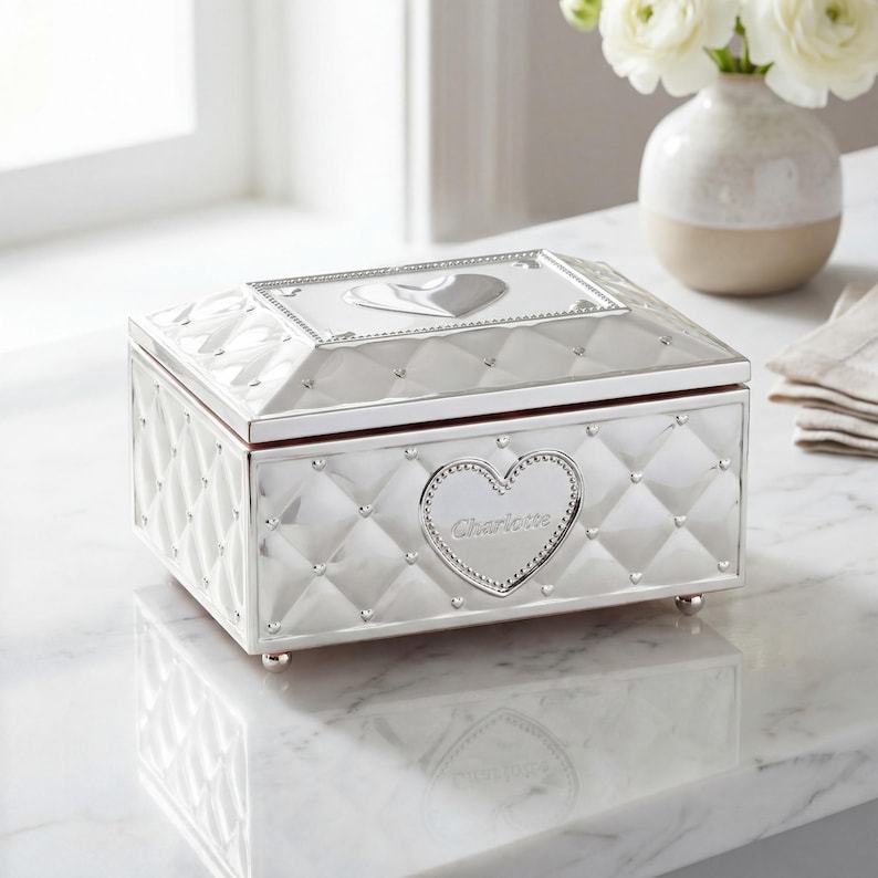 May include: A silver jewellery box with a quilted design and heart-shaped details. The box has a heart-shaped emblem with the name "Charlotte" and rests on small, rounded feet. A decorative item for storing jewellery.