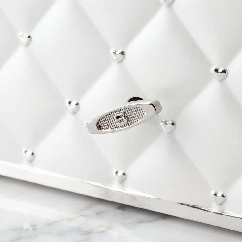 May include: Close-up of a white, quilted jewelry box with silver heart-shaped studs. The box features a silver handle with a textured design. The box has a silver trim and a white interior.