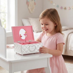 May include: A white, quilted jewellery box with a pink interior and a heart-shaped emblem. The box features a mirror and a ballerina figurine. The box is open, revealing compartments for jewellery. The box is on a white table.