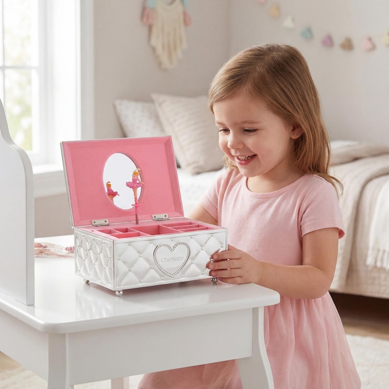 May include: A white, quilted jewellery box with a pink interior and a heart-shaped emblem. The box features a mirror and a ballerina figurine. The box is open, revealing compartments for jewellery. The box is on a white table.