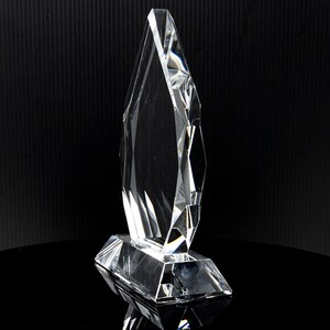 Puede incluir: Un premio de cristal transparente con un dise&ntilde;o en forma de diamante. El premio tiene una base y una parte superior alta y puntiaguda.