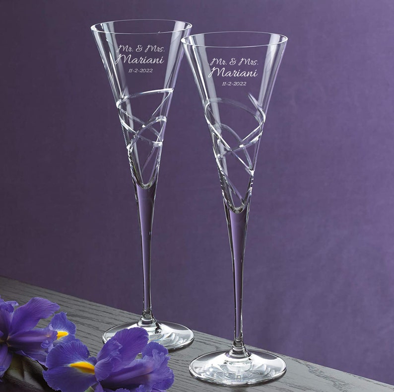 May include: Two clear glass champagne flutes with a swirling design. The flutes are engraved with the text "Mr. & Mrs. Mariani" and the date "11-2-2022".