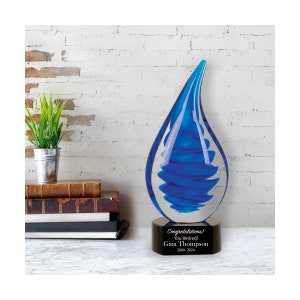 May include: Blue glass teardrop award with a black base. The base has the text "Congratulations! You Retired! Gina Thompson 2000-2024".