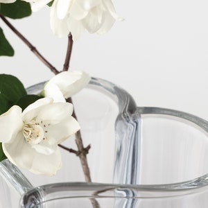 Op de afbeelding: Een heldere glazen vaas in hartvorm, met een takje witte jasmijnbloemen met groene bladeren. De bloemen staan in volle bloei, met delicate bloemblaadjes en gele centra. De vaas staat op een witte achtergrond.
