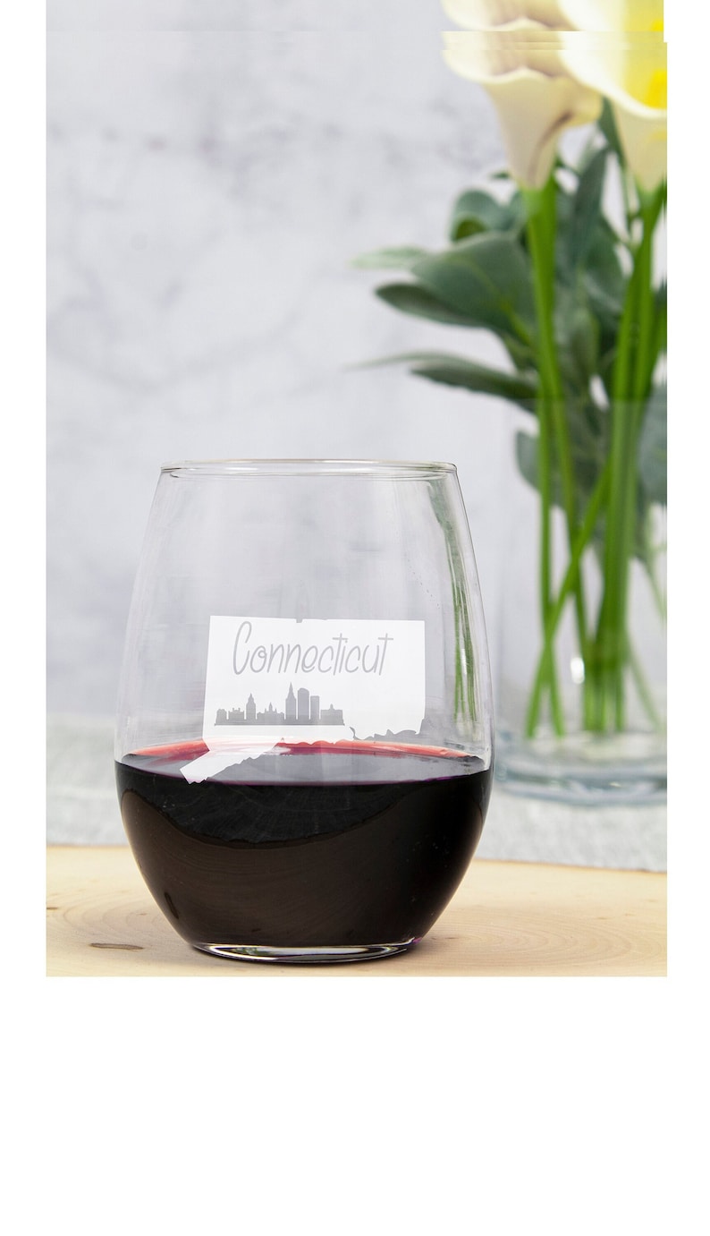 May include: A clear glass wine glass filled with red wine. The glass features a white outline of the state of Connecticut with the word "Connecticut" written on it.