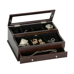 May include: A dark brown wooden jewelry box with a glass top, open to reveal compartments lined with black fabric. The box contains a watch, rings, necklaces, and money. A drawer is partially open, displaying a necklace. The box has silver knobs.