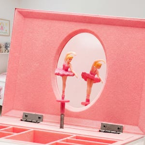 May include: A pink jewellery box featuring a ballerina figurine. The box has an oval mirror window with a ballerina in a pink tutu. The ballerina stands on a pink stand. The box is open, showing compartments.
