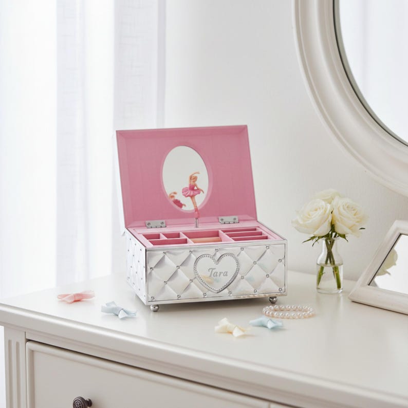 May include: Silver jewellery box with a pink interior and a ballerina design. The box has a quilted pattern, a heart-shaped nameplate, and a mirror inside the lid. The box is open, revealing compartments for jewellery.