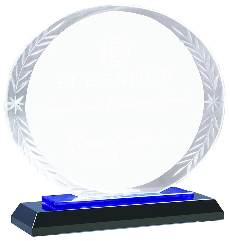 May include: A clear glass award with a blue base. The award has a circular design with a laurel wreath etched around the edge. The text "Elegance" is engraved on the award.