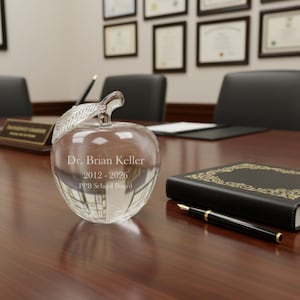 Puede incluir: Un premio transparente en forma de manzana de cristal con un tallo de hoja, grabado con "Dr. Brian Keller 2012-2026 PPB School Board". El premio est&aacute; sobre una mesa de madera junto a un libro negro y un bol&iacute;grafo.
