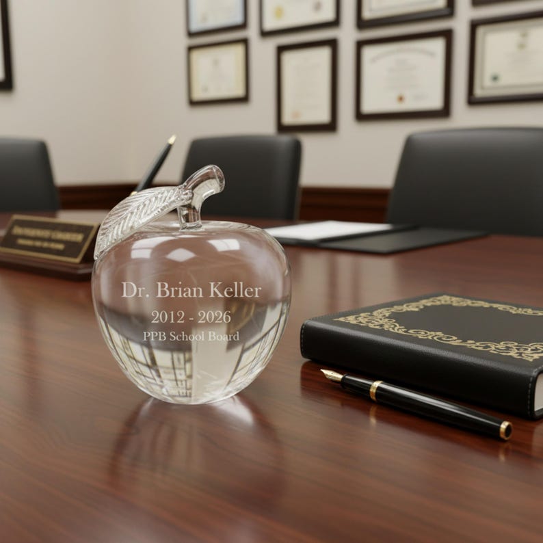 Puede incluir: Un premio transparente en forma de manzana de cristal con un tallo de hoja, grabado con "Dr. Brian Keller 2012-2026 PPB School Board". El premio est&aacute; sobre una mesa de madera junto a un libro negro y un bol&iacute;grafo.