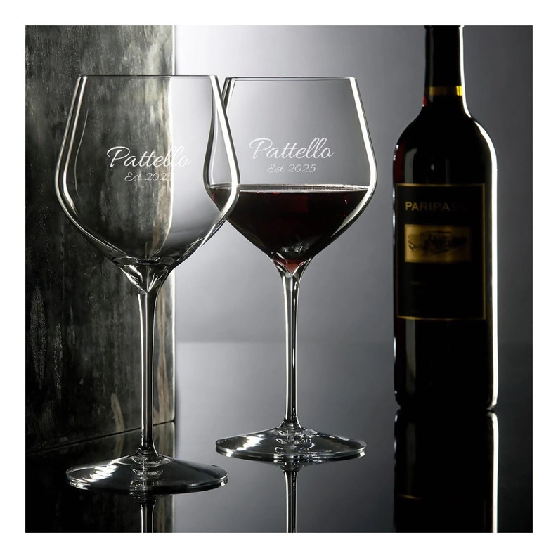 May include: Two clear wine glasses with the text "Pattello Est 2025" etched on the side. One glass is filled with red wine. A bottle of red wine is in the background.
