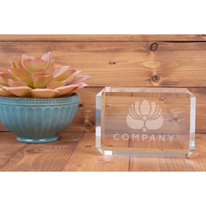 May include: A clear glass paperweight with a frosted design of a lotus flower and the word "COMPANY" engraved on the bottom. The paperweight is sitting on a wooden surface.