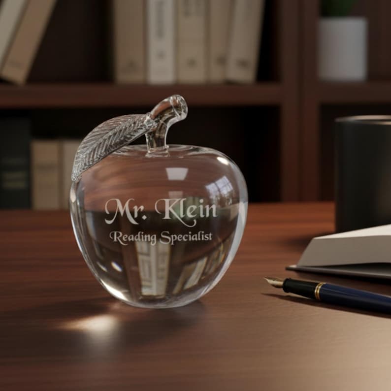 Puede incluir: Un pisapapeles transparente en forma de manzana con un asa en forma de hoja. El texto "Mr. Klein Reading Specialist" est&aacute; grabado en la parte delantera. Un bol&iacute;grafo y un libro abierto est&aacute;n sobre el escritorio de madera.