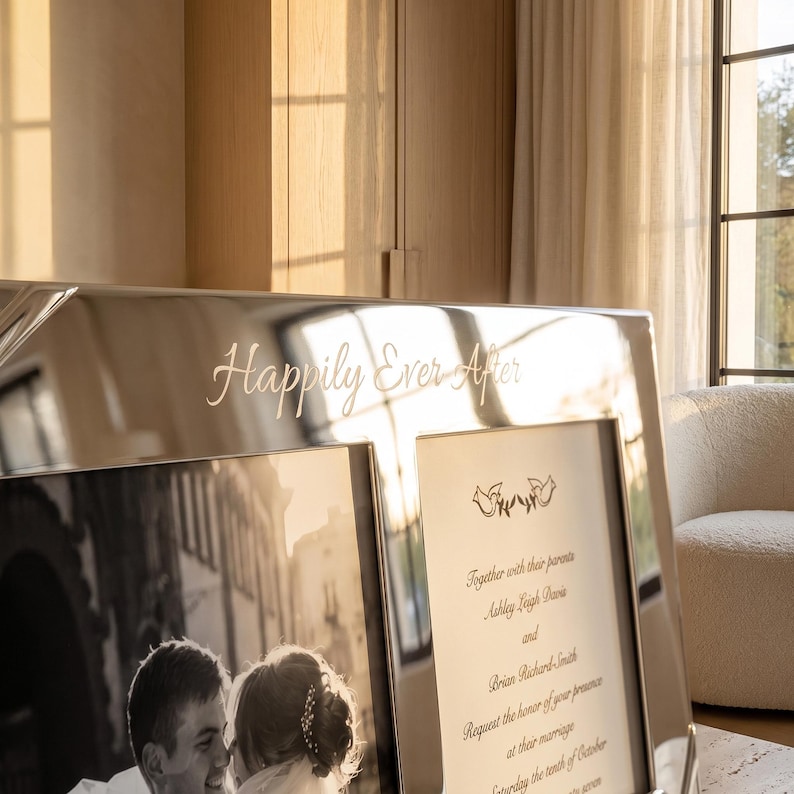 May include: A silver-toned metal double picture frame featuring the engraved text "Happily Ever After" at the top. One side holds a black and white photograph of a couple, while the other side displays a printed wedding invitation with two birds.