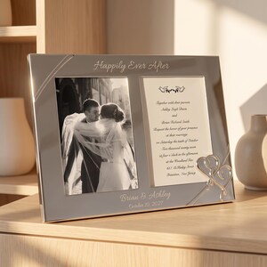 May include: A silver-toned double photo frame featuring the text "Happily Ever After" at the top and "Brian & Ashley October 10, 2027" at the bottom. The left side holds a black and white photo of a couple, while the right side displays wedding invitation text with two small bird icons and a decorative silver heart accent.