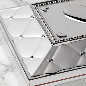 May include: A close-up of a silver box with a diamond pattern and heart-shaped details. The box has a reflective surface and a decorative border with small, sparkling embellishments. The design is elegant and detailed.