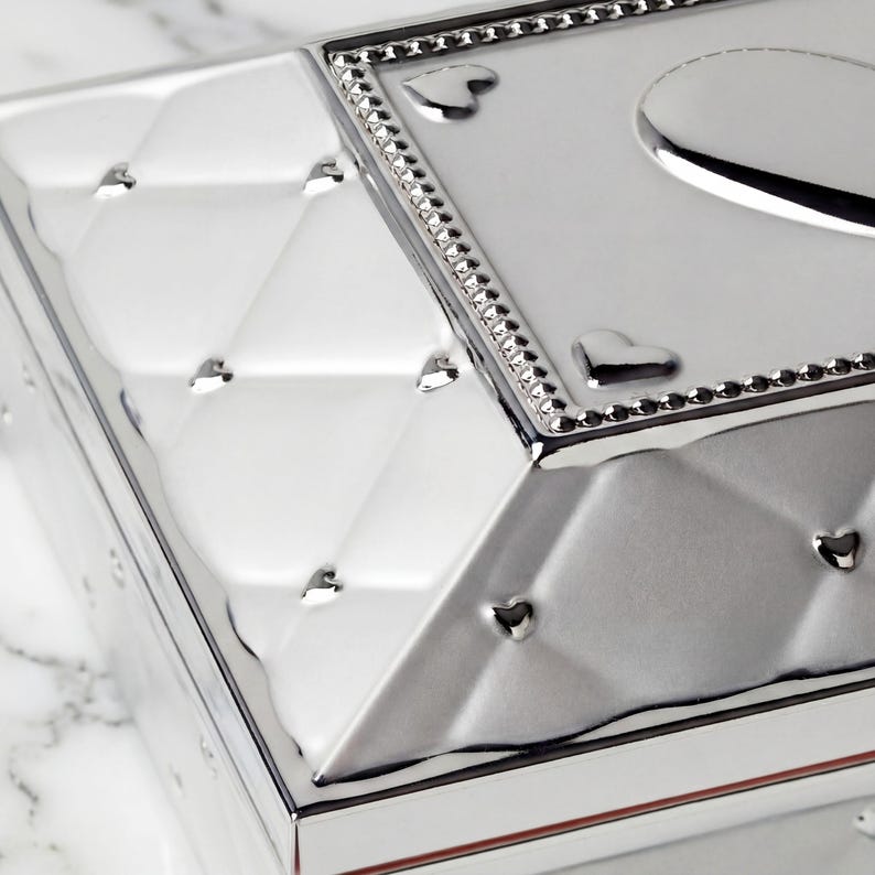 May include: A close-up of a silver box with a diamond pattern and heart-shaped details. The box has a reflective surface and a decorative border with small, sparkling embellishments. The design is elegant and detailed.