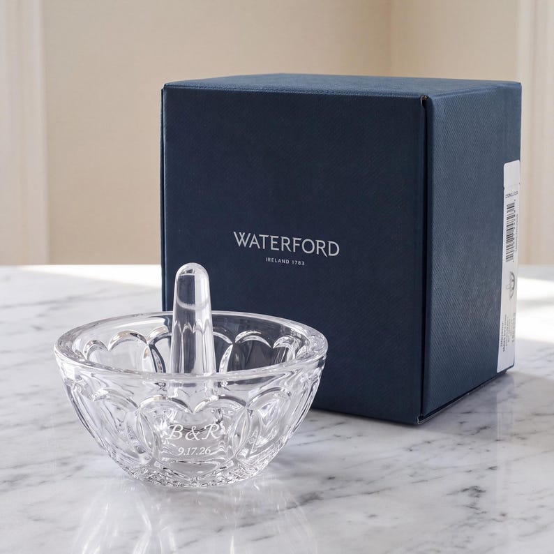 May include: A clear crystal ring holder with a central post and a scalloped bowl design. The bowl is etched with the letters B&R and the date 9.17.26. It is positioned in front of a dark blue square box with the text WATERFORD IRELAND 1783.