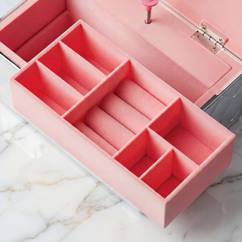 May include: Open pink jewellery box with multiple compartments and ring slots. The interior is lined with soft pink fabric. The exterior is silver with a quilted pattern. A pink knob is visible on the inside.