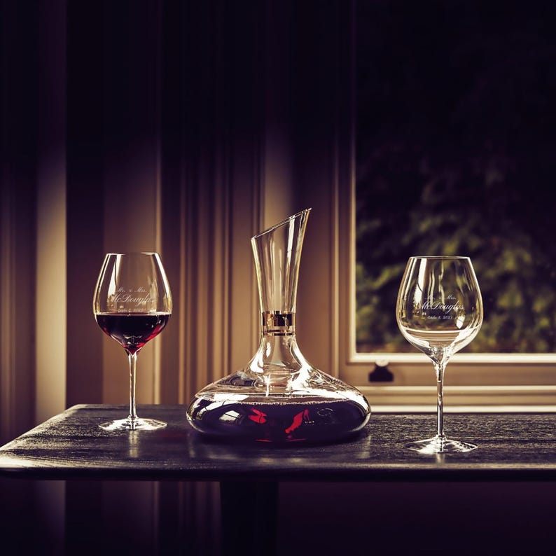 May include: A clear glass decanter with a wide base and a long neck, filled with red wine. Two clear wine glasses with engraved text are on either side of the decanter, one filled with red wine and the other empty. The decanter and glasses are on a dark wooden table.