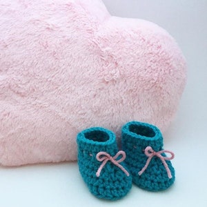 May include: A pair of teal blue crocheted baby booties with pink ribbon ties. The booties are sitting on a pink fluffy heart-shaped pillow.