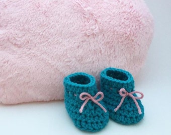 Op de afbeelding: Een paar babyblauwe gehaakte babyschoentjes met roze lintjes. De schoentjes liggen op een roze, pluche hartvormig kussen.