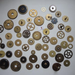 Op de afbeelding: Een verzameling vintage horlogetandwielen in messing en zwart, in verschillende maten. De tandwielen zijn gerangschikt op een witte ondergrond. Sommige tandwielen hebben zichtbare tanden, andere spaken of massieve centra. Een klein wit label met het nummer "3" is ook aanwezig.