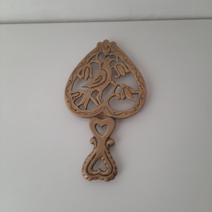 May include: A gold-coloured metal trivet in the shape of a heart with a bird and floral design. The trivet has a handle in the shape of a heart.