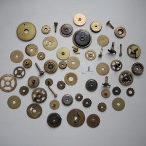 Op de afbeelding: Een verzameling vintage horlogetandwielen en -componenten in verschillende maten en vormen, inclusief schroeven en rondsel. De tandwielen zijn voornamelijk messingkleurig, sommige vertonen tekenen van ouderdom en slijtage. Er is ook een kleine witte kaart met het nummer "1".