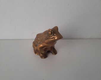 Pode incluir: Estatueta de rã em bronze. A rã está sentada, com textura detalhada nas costas e na cabeça. A estatueta tem um brilho metálico e está sobre um fundo liso.