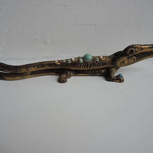 May include: A bronze alligator sculpture with turquoise and gold accents. The alligator has a detailed design with open jaws and is decorated with small, round turquoise and gold-colored stones. The sculpture is placed on a white surface.