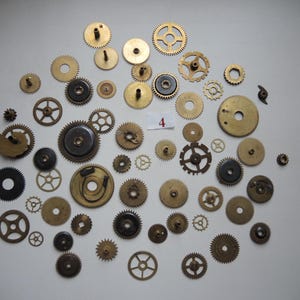 Op de afbeelding: Een verzameling van verschillende maten messing en donker metalen horlogetandwielen verspreid op een witte ondergrond. Sommige tandwielen hebben tanden, andere hebben een glad, cirkelvormig ontwerp. Een klein wit label met het nummer "4" is ook aanwezig.