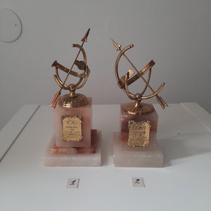 May include: Two gold-colored trophies with a globe and arrow design on top of marble bases. The bases are inscribed with the text "Rakna de Lyckliga" and "Glada Gaster", respectively. The trophies are numbered 1 and 2.