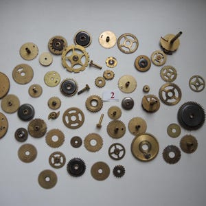 Op de afbeelding: Een verzameling messing en donker metalen tandwielen, raderen en schroeven van verschillende groottes, verspreid over een wit oppervlak. De tandwielen hebben verschillende aantallen tanden en patroon gaten. Sommige tandwielen zijn cirkelvormig, andere hebben spaken.